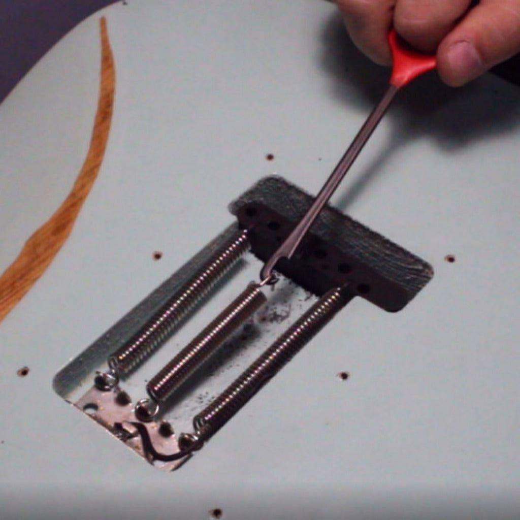 A close-up of a guitar tailpiece being adjusted with a red-handled screwdriver, mounted on a white electric guitar body.