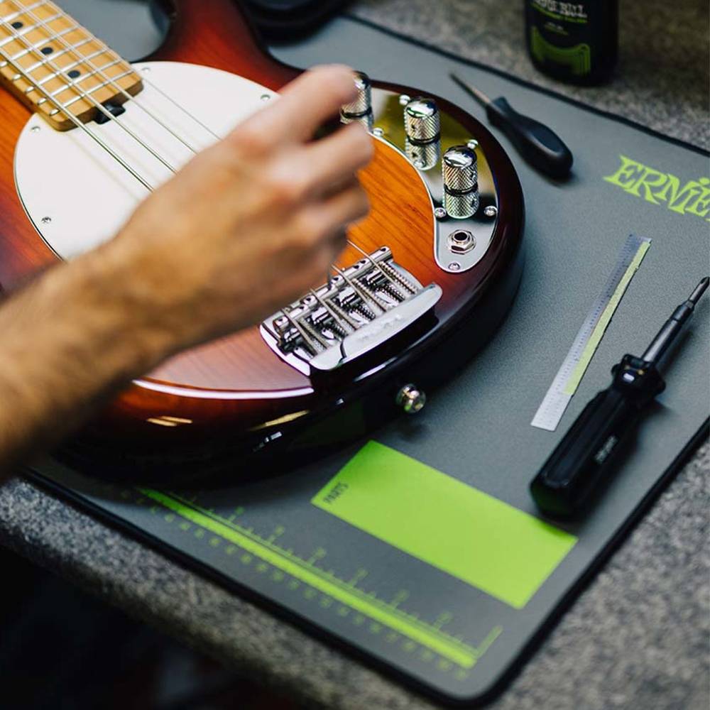 A close-up of a person's hand adjusting the tuning pegs of a brown electric bass guitar on a dark gray work mat with green markings. A black screwdriver, a metal file, and other tools are arranged on the mat on a gray workbench.