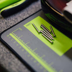 Close-up of a black and neon green parts measurement mat with several metal screws placed on it, photographed on a textured gray surface.