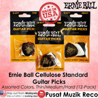Three packages of Ernie Ball Cellulose Standard Guitar Picks in Thin, Medium, and Heavy sizes, shown in assorted black, white, and brown colors. The image features a pink background with text overlays detailing the product, including 'Assorted Colors, Thin/Medium/Hard (12-Pack)' and a 'Made in U.