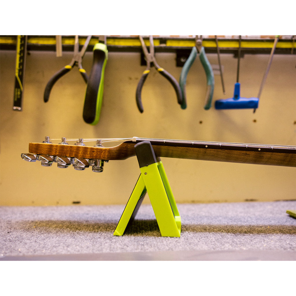 A guitar headstock and neck resting on a bright neon yellow A-frame stand in a workshop setting, with hanging tools visible in the background.