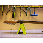 A guitar headstock and neck resting on a bright neon yellow A-frame stand in a workshop setting, with hanging tools visible in the background.