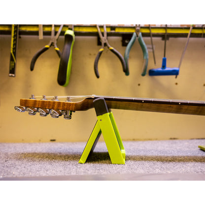 A guitar headstock and neck resting on a bright neon yellow A-frame stand in a workshop setting, with hanging tools visible in the background.