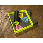 A black cylindrical guitar pick holder with a neon green frame, shown on a dark gray insert holding three colorful guitar picks (yellow, blue, and pink) on a wooden tabletop.