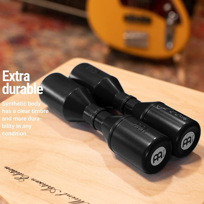 Two black Meinl acoustic guitar string tree accessories with white Meinl logos, resting on a light wood cutting board with a blurred yellow guitar in the background. White text on the left reads 'Extra durable' and 'Synthetic body has a clear timbre and more durability in any condition.
