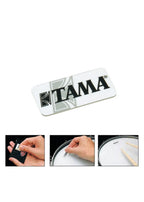 TAMA logo on a white background with a hand holding a small black card and a drum pad.