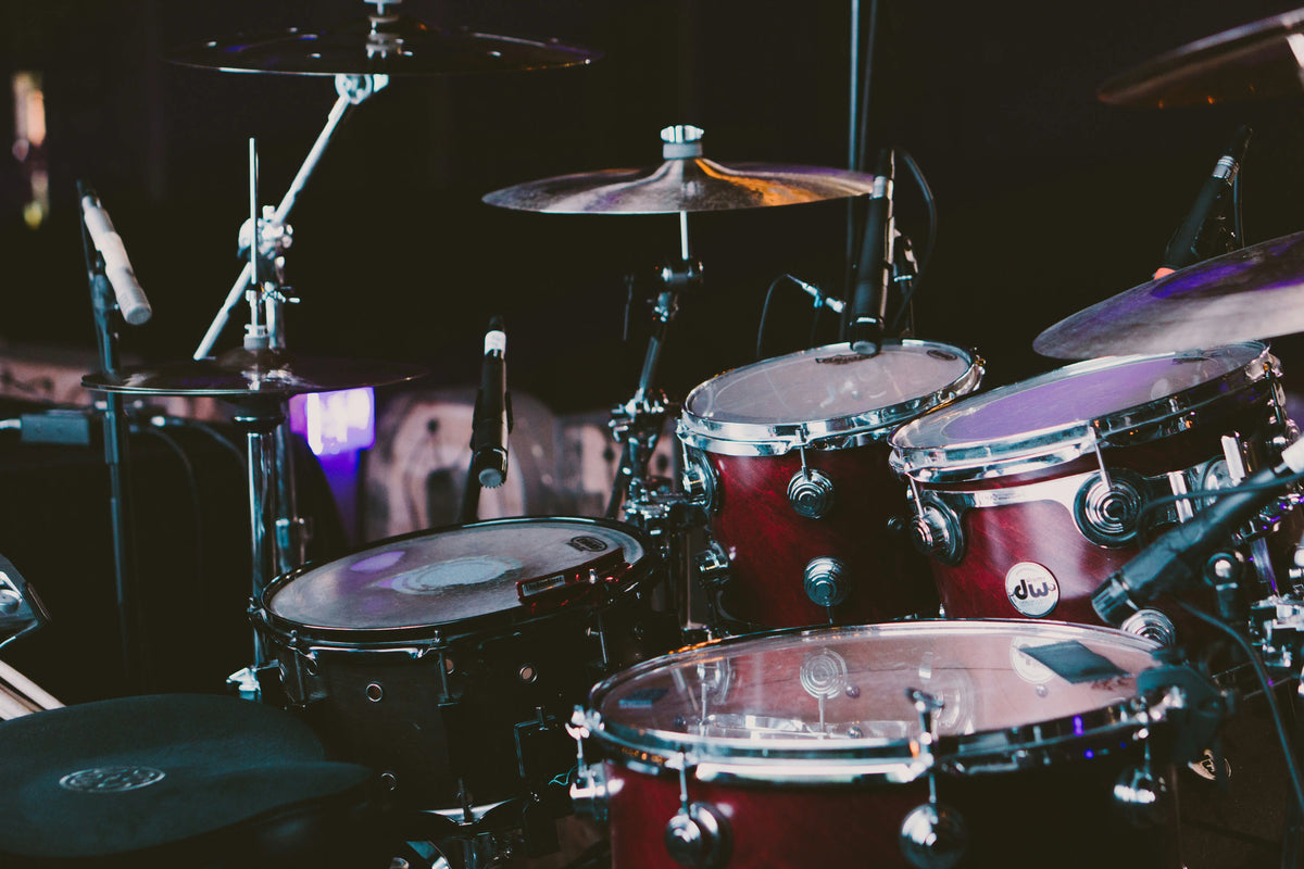 A close-up, slightly moody shot of a drum kit with red and black shells, chrome hardware, and cymbals on stands in a dimly lit setting.